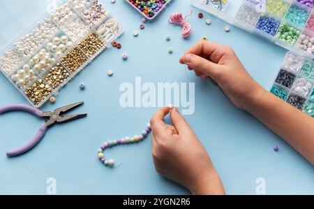 Una persona infiltra abilmente perline su una corda, circondata da vari contenitori di perline colorate e strumenti su un luminoso tavolo da lavoro. Foto Stock