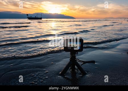Action camera con treppiede per riprese estive in spiaggia. Riprese di un video temporizzato su action camera al tramonto o all'alba sulla spiaggia. Montagne, crepuscolo. Trave Foto Stock