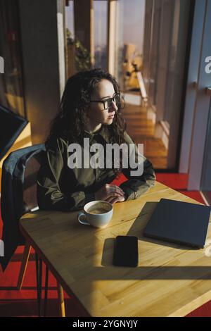 Una donna carina lavora su un notebook in un bar. Giovane donna concentrata bruna in bicchieri seduta al tavolo vicino alla finestra a bere caffè. Freelance Foto Stock