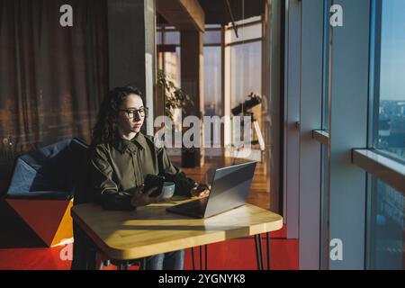 Una donna carina lavora su un notebook in un bar. Giovane donna concentrata bruna in bicchieri seduta al tavolo vicino alla finestra a bere caffè. Freelance Foto Stock