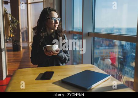 Una donna carina lavora su un notebook in un bar. Giovane donna concentrata bruna in bicchieri seduta al tavolo vicino alla finestra a bere caffè. Freelance Foto Stock