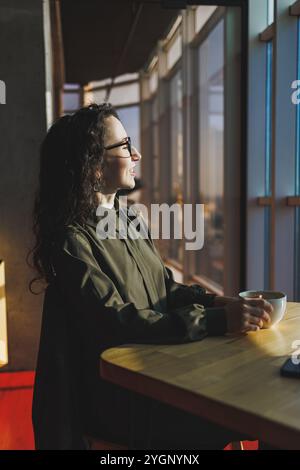 Una donna carina lavora su un notebook in un bar. Giovane donna concentrata bruna in bicchieri seduta al tavolo vicino alla finestra a bere caffè. Freelance Foto Stock