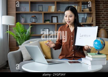 Donna asiatica che conduce lezioni online da casa, tenendo una lavagna con la lezione scritta su di essa, utilizzando computer portatile e telefono. La scena raffigura l'apprendimento a distanza, la comunicazione e l'istruzione moderna. Foto Stock