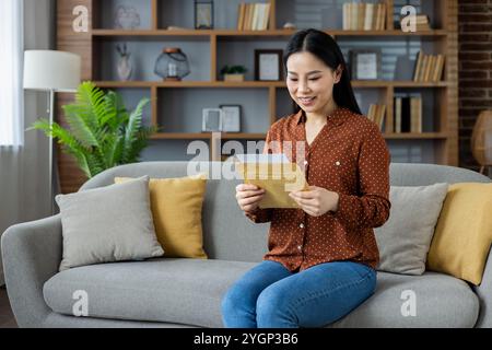 Una donna asiatica gioiosa seduta sul divano legge la lettera con gioia. Abbigliamento casual, ambiente rilassato. La scena cattura l'emozione, il relax e la connessione in ambiente domestico. Foto Stock