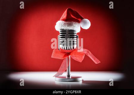 Microfono retrò per prestazioni elevate con illuminazione spot decorato con cappello di Natale e fiocco rosso su tavolo bianco isolato su sfondo rosso. Anteriore Foto Stock