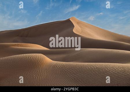 Duna di sabbia dorata a le Khwair, Oman, circondata da motivi di sabbia liscia e ondulati, sotto un cielo blu brillante con nuvole sparse. Foto Stock