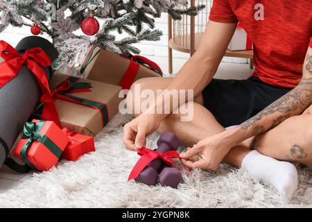 Giovane uomo tatuato che disimballa il regalo dei manubri sul pavimento vicino all'albero di Natale Foto Stock