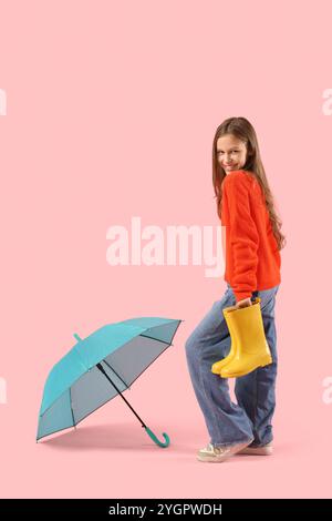 Simpatica ragazza adolescente con stivali da pioggia in gomma e ombrello blu su sfondo rosa Foto Stock