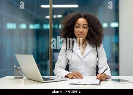 Donna afroamericana dottore in carica. È seduta alla scrivania con un computer portatile, concentrata sui documenti. L'immagine ritrae professionalità, attenzione e fiducia in un ambiente medico moderno. Foto Stock