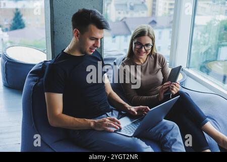 Gli studenti si siedono nell'area di lavoro vicino alla finestra e lavorano insieme su un progetto su un notebook. I colleghi allegri manager guardano il laptop insieme e le lacune Foto Stock