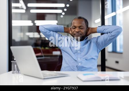 Uomo d'affari afroamericano maturo che si rilassa alla scrivania dell'ufficio, mani dietro la testa, laptop aperto. Indossa una camicia blu che esprime sollievo e soddisfazione. Spazio di lavoro moderno, atmosfera di lavoro senza stress. Foto Stock