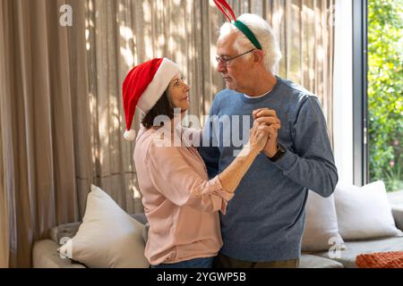 Natale, coppia senior che balla a casa, indossa cappelli festivi e sorride calorosamente. Celebrazione, anziani, danza, felicità, festa, insieme Foto Stock