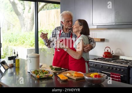 Una coppia di anziani che cucinano insieme in cucina, condividendo momenti di amore con il vino, a casa Foto Stock