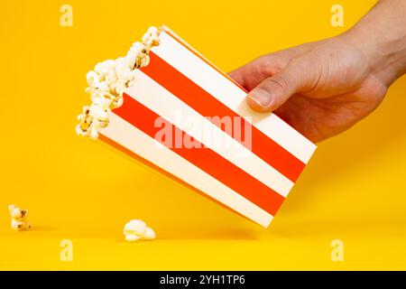 Mano maschio che tiene un secchio di cartone rosso e bianco con popcorn su sfondo giallo, concetto di popcorn Foto Stock