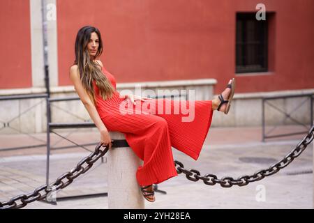 Una donna elegante vestita con uno splendido abito rosso che si posa elegantemente su una struttura a catena Foto Stock