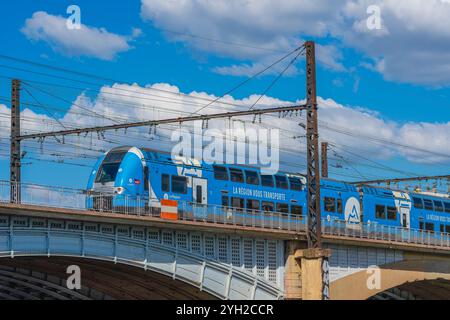 Lione, Francia. 24/06/12A il treno blu SNCF attraversa un ponte sotto un cielo limpido durante il giorno, circondato da nuvole sparse e infrastrutture ferroviarie Foto Stock