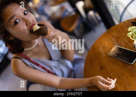 Una giovane donna allegra che assaggia un cono di gelato al cioccolato in un'atmosfera rilassata da caffetteria, catturando un momento giocoso e spensierato. Foto Stock