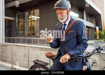 Un vecchio uomo d'affari barbuto con casco e bicicletta che invia SMS sullo smartphone mentre si sposta per andare al lavoro in città Foto Stock