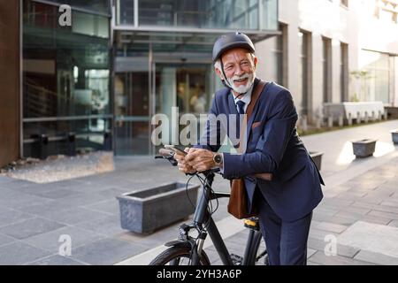 Allegro professionista maschile con casco che usa il telefono e sorride mentre si appoggia in bicicletta sulla strada della città Foto Stock