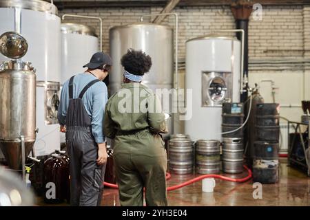 Vista posteriore di un team di due operai della fabbrica di sidro che ispezionano il processo di produzione sul pavimento dell'officina con enormi serbatoi di fermentazione presso l'impianto, copia spa Foto Stock