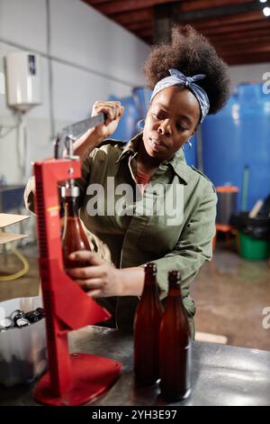 Shot verticale di una donna afroamericana che lavora sodo, tappando bottiglie di vetro riempite con sidro artigianale o birra utilizzando l'attrezzo manuale a f Foto Stock
