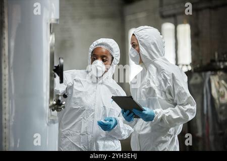 Due tecnici di processo donne professionisti che indossano maschere e tute hazmat che ispezionano le apparecchiature in un'officina produttrice di sidro, mentre collaborano contro Foto Stock