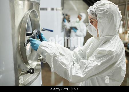 Vista laterale del tecnico di processo donna che indossa tuta antimanomissione e guanti protettivi, che attiva la valvola sul serbatoio riempito di liquido durante l'ispezione del ferm del sidro Foto Stock