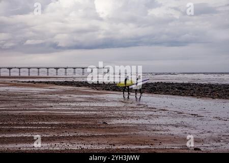 Due surfisti che camminano intenzionalmente attraverso le sabbie di Saltburn Beach con il molo sullo sfondo, Saltburn, Teesside, Inghilterra. Foto Stock