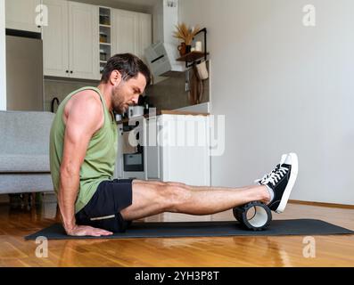 Persona maschile che esegue esercizi di rilascio muscolare su un tappetino da yoga con un rullo in schiuma a casa. Foto Stock