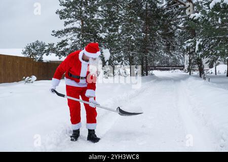 Babbo Natale pulisce la neve con la pala in inverno all'aperto dopo una nevicata. Pulire le strade del villaggio, pulire il passaggio per le auto, difficile Foto Stock
