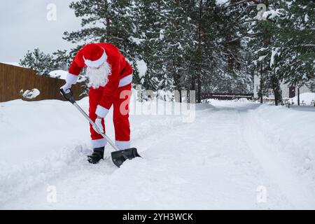 Babbo Natale pulisce la neve con la pala in inverno all'aperto dopo una nevicata. Pulire le strade del villaggio, pulire il passaggio per le auto, difficile Foto Stock