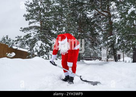 Babbo Natale pulisce la neve con la pala in inverno all'aperto dopo una nevicata. Pulire le strade del villaggio, pulire il passaggio per le auto, difficile Foto Stock