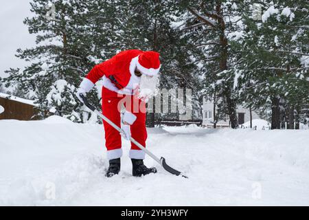 Babbo Natale pulisce la neve con la pala in inverno all'aperto dopo una nevicata. Pulire le strade del villaggio, pulire il passaggio per le auto, difficile Foto Stock