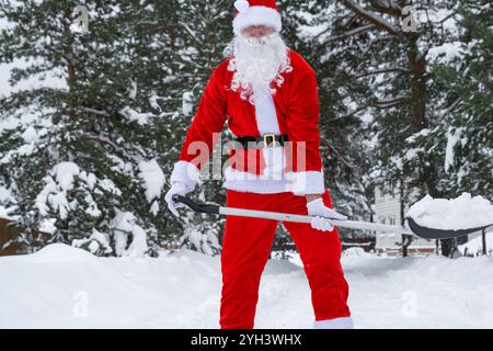 Babbo Natale pulisce la neve con la pala in inverno all'aperto dopo una nevicata. Pulire le strade del villaggio, pulire il passaggio per le auto, difficile Foto Stock
