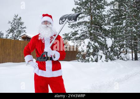 Babbo Natale pulisce la neve con la pala in inverno all'aperto dopo una nevicata. Pulire le strade del villaggio, pulire il passaggio per le auto, difficile Foto Stock
