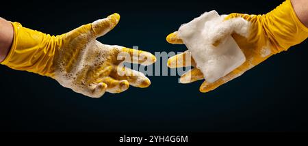 Due mani in guanti di gomma gialli. Una mano tiene una spugna nella schiuma e l'altra la tiene. Il processo di lavaggio dei piatti o di pulizia. Foto Stock