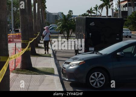 Vista da dietro una donna anziana che cammina lungo le linee principali del marciapiede con nastro adesivo giallo, auto, rimorchio della spazzatura e palme. Spiaggia sabbiosa sulla sinistra di Gulfp Foto Stock