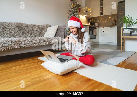 Una donna con il cappello di Babbo Natale siede sul pavimento in posa di loto, usando il laptop per videochiamate e gesti con le mani del cuore. Foto Stock