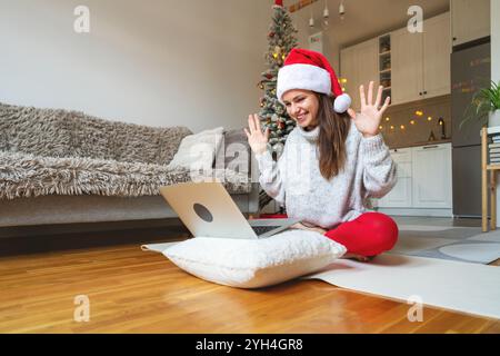 Giovane donna con un cappello di Babbo Natale seduta in posa di loto, videochiama gli amici sul portatile, sorride e saluta. Foto Stock