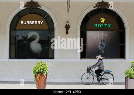 Hanoi, Vietnam - 10 luglio 2023: Una donna vietnamita, che indossa il tradizionale cappello non la, corre in bicicletta davanti a un negozio Burberry e Rolex. Foto Stock