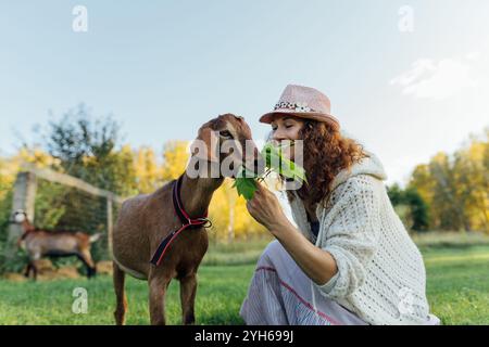 Una donna dà da mangiare a una capra con foglie fresche mentre è seduta in una fattoria in campagna Una donna contenta si impegna con una capra, simboleggiando la pacifica a Foto Stock