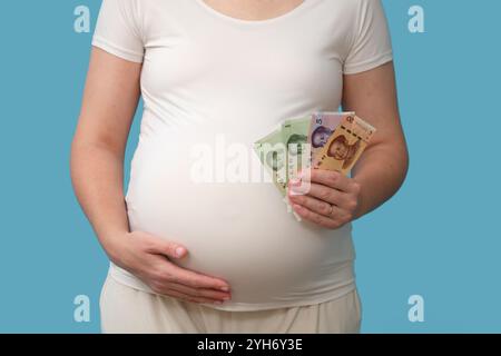 Banconote in yuan nelle mani di una donna durante la gravidanza, isolate su uno sfondo blu studio. Una donna incinta tiene soldi cinesi yuan, concetto Foto Stock