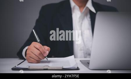 Uomo d'affari in abbigliamento formale che firma i documenti a una scrivania con un computer portatile, concentrandosi sulla documentazione e sul completamento del contratto in un ufficio. Foto Stock