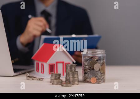 Uomo d'affari in background che esamina documenti con un modello di casa, pile di monete e barattoli di monete sul tavolo, che rappresentano la pianificazione finanziaria e. Foto Stock