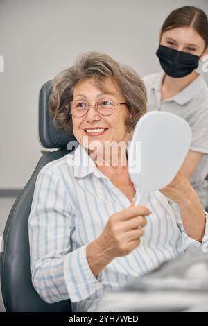 Signora dai capelli grigi in studio dentistico con specchio in mano Foto Stock