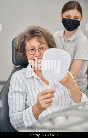 La signora dai capelli grigi esamina i suoi denti nello specchio Foto Stock