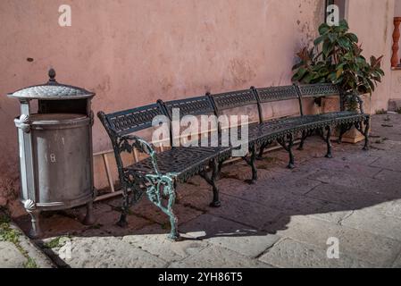 Una lunga panchina di metallo per cinque persone, un cestino sulla strada asfaltata di Porto Azzurro nell'isola d'Elba in Italia Foto Stock