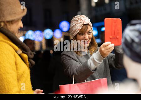 Donna elegante con fasciatesta turbante che controlla il suo riflesso nello specchio portatile mentre fa shopping durante le feste serali Foto Stock
