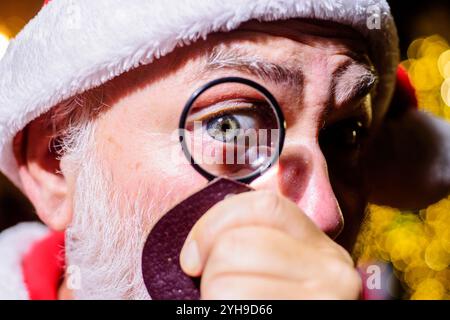 Vacanze di Natale o Capodanno. Babbo Natale guarda attraverso una lente d'ingrandimento. Babbo Natale guarda con curiosità attraverso la lente d'ingrandimento su di te. Barbuti Foto Stock