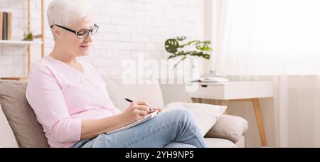 Signora senior che scrive la lista di shopping a casa, panorama Foto Stock